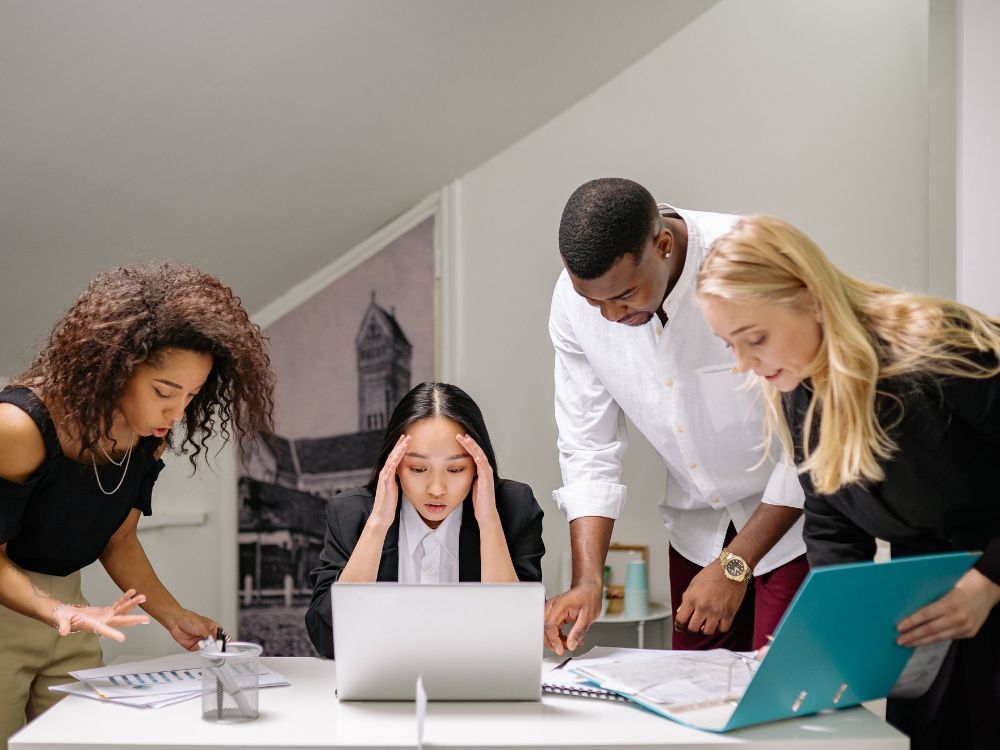 Four colleagues crowded around a stressed coworker at a desk piled with printouts and folders